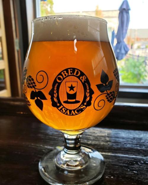 A glass of golden beer with a logo reading "Obed & Isaac's" sits on a wooden table, with a blurred background.