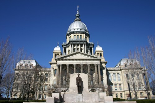 The Illinois State Capitol building with a statue of Abraham Lincoln in the foreground against a clear blue sky.