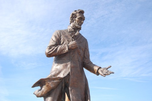 A bronze statue of Abraham Lincoln, standing with one hand extended, against a blue sky.