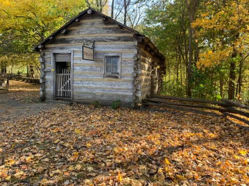 A rustic log cabin surrounded by colorful autumn leaves and trees, with a wooden fence in the foreground.