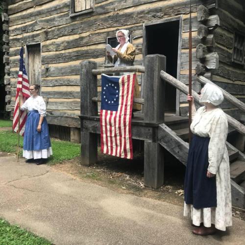 Three women in historical attire stand outside a log cabin, one reading from a document, with a flag displayed.