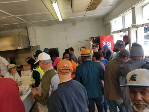 A busy restaurant scene with a crowd of people waiting in line, some interacting with staff at a serving counter.