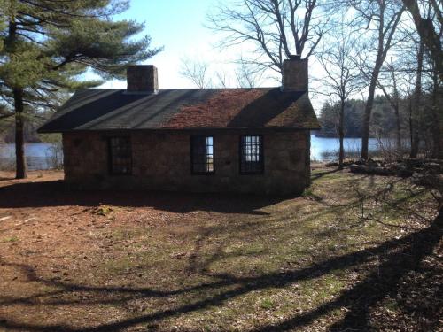 A stone cottage by a lake, surrounded by trees and sunlight, with a clear blue sky in the background.