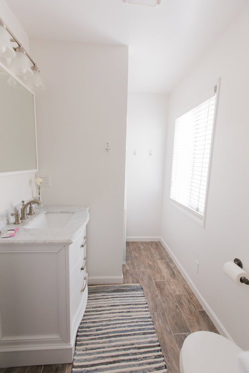 Bright, modern bathroom featuring a white vanity, mirror, and a window with blinds, complemented by a striped rug.