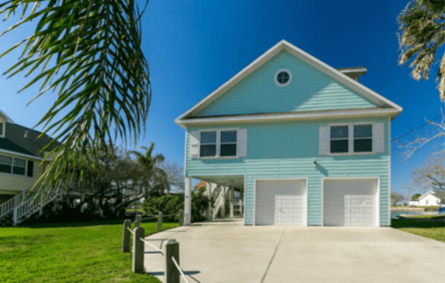 A light blue house with a garage, surrounded by greenery and palm trees, under a clear blue sky.