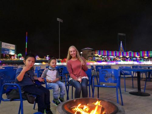 Two children and a teenager sit by a fire pit, roasting marshmallows, with colorful lights in the background at night.