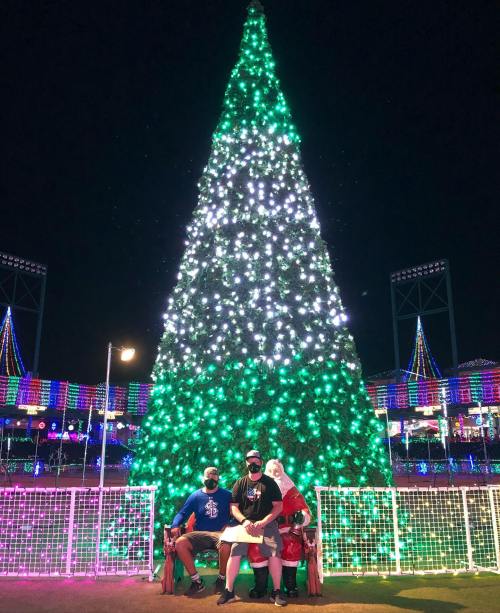 A large, brightly lit Christmas tree with two people sitting on a bench, one dressed as Santa, at a festive night event.
