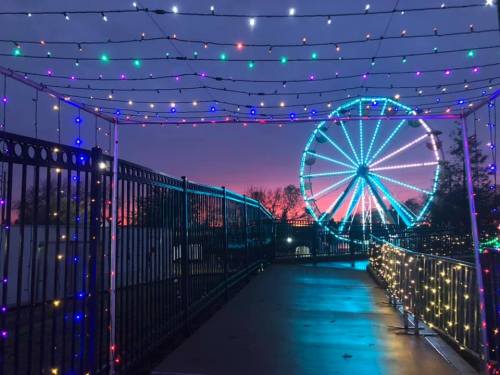 A colorful ferris wheel illuminated by lights at dusk, framed by a pathway adorned with festive string lights.