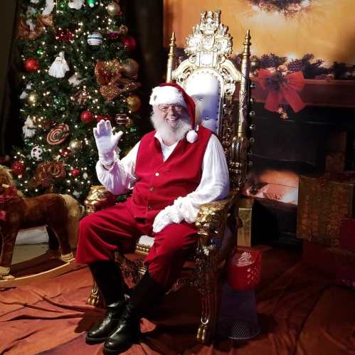 Santa Claus in a red suit sits on a golden throne, waving, with a decorated Christmas tree in the background.