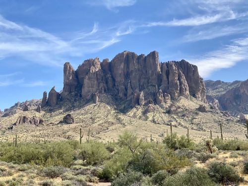 A rugged mountain range with steep cliffs and desert vegetation under a blue sky with wispy clouds.