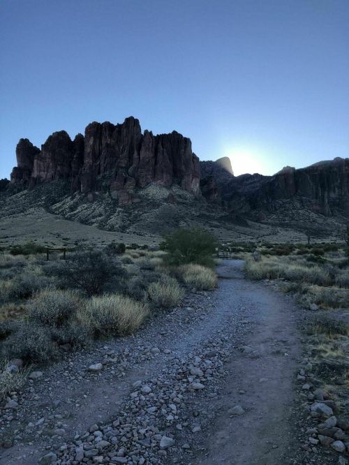 A rocky path leads towards a mountain range at dawn, with the sun rising behind the peaks in a clear blue sky.