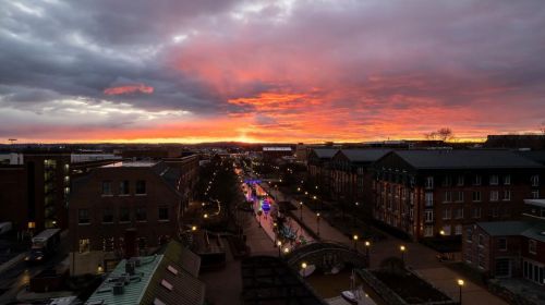 A vibrant sunset casts orange and pink hues over a cityscape, with streets illuminated by lights and clouds above.