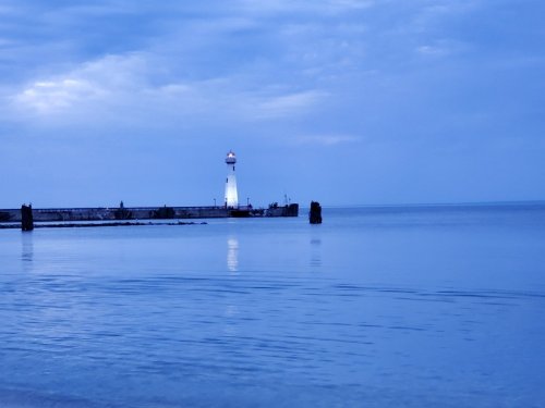 A calm seascape at dusk featuring a lighthouse on a pier, reflecting in the still water.