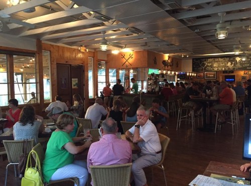 A busy restaurant interior with patrons dining and socializing at tables and the bar, featuring wooden decor and bright lighting.
