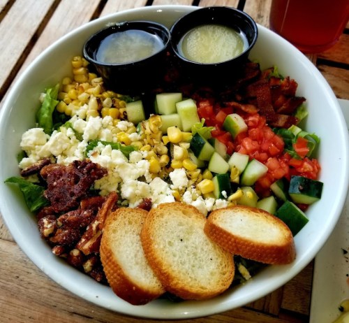 A colorful salad with greens, corn, tomatoes, cucumbers, bacon, cheese, and two dressings, served with toasted bread.