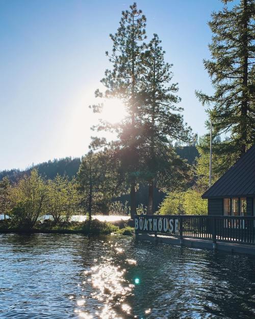 Sunlight glimmers on water near a boathouse, surrounded by trees and mountains under a clear blue sky.
