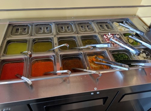 A condiment bar with various sauces and toppings in metal containers, some partially filled, with tongs for serving.