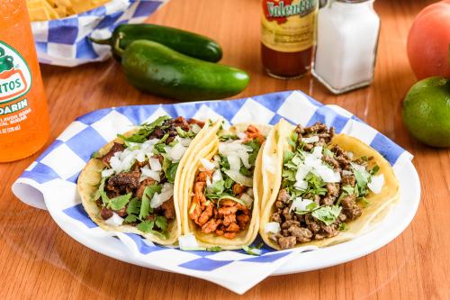 A plate of three tacos filled with meat, topped with cilantro and onions, served with drinks and condiments in the background.