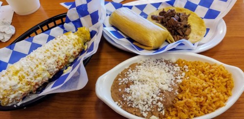 A plate of Mexican food featuring a tamale, taco with beef, and sides of refried beans and rice, all on checkered paper.