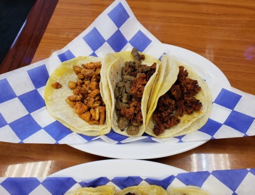 Three tacos on a plate: one with chicken, one with chorizo, and one with beef, served on blue and white checkered paper.
