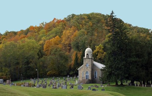 A stone church surrounded by a cemetery, with colorful autumn trees on a hillside in the background.