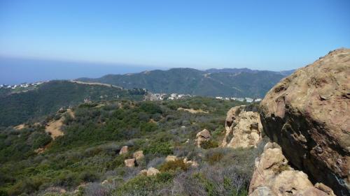A panoramic view of rolling green hills and rocky terrain under a clear blue sky, with distant mountains and a coastline.