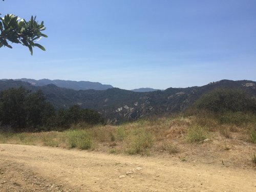 A scenic view of rolling hills and mountains under a clear blue sky, with a dirt path in the foreground.