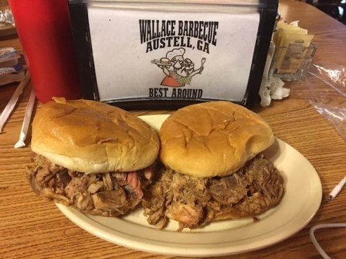 Two pulled pork sandwiches on a plate, with a sign for Wallace Barbecue in the background.