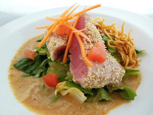 A plate of sesame-crusted tuna on a bed of lettuce, topped with carrots, tomatoes, and crispy noodles in a sauce.