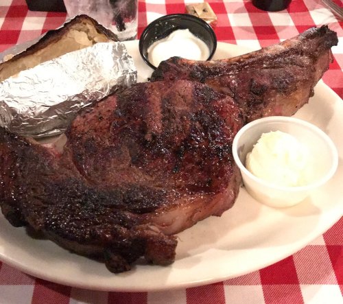A large, grilled ribeye steak served with a baked potato and a side of butter on a checkered tablecloth.