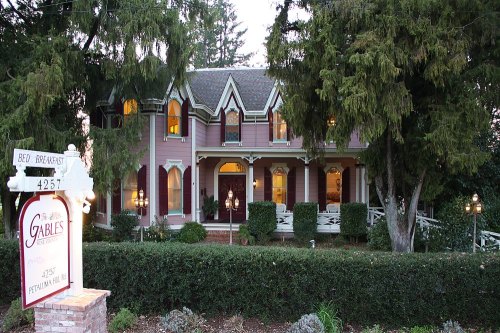A charming pink Victorian bed and breakfast surrounded by trees and greenery, with a welcoming sign in front.