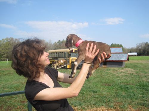 A woman holds a small brown puppy in a field, with a school bus and barn in the background under a blue sky.