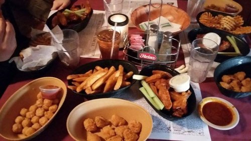 A table filled with various dishes: chicken wings, fries, tater tots, celery, and dipping sauces, with drinks nearby.