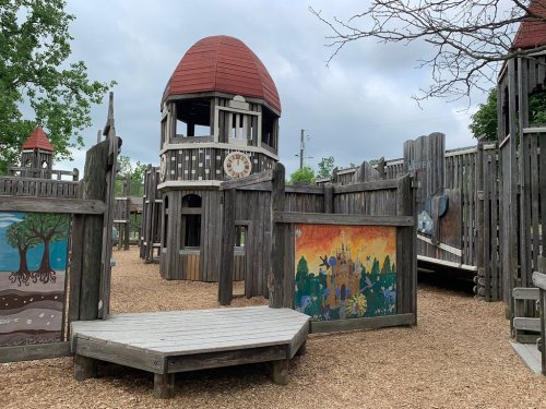 A wooden playground with a tower, red roofs, and colorful murals, surrounded by gravel and trees under a cloudy sky.
