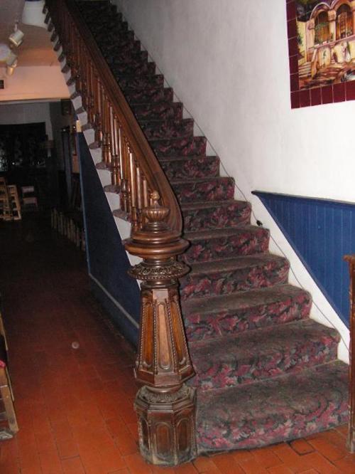A wooden staircase with a floral carpet, featuring ornate banisters and a decorative post at the bottom.