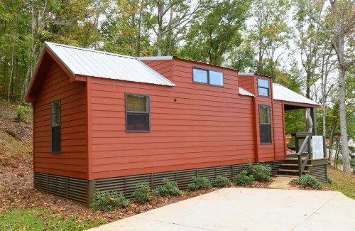A modern red cabin with a metal roof, surrounded by trees and a concrete driveway.