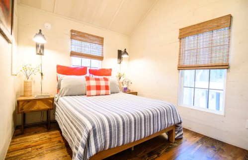 Cozy bedroom with a striped bedspread, red and gray pillows, wooden nightstand, and natural light from a window.