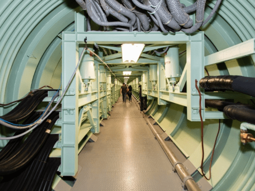 A long, narrow corridor inside a green-tinted tunnel with cables and pipes along the walls. Two people walk in the distance.