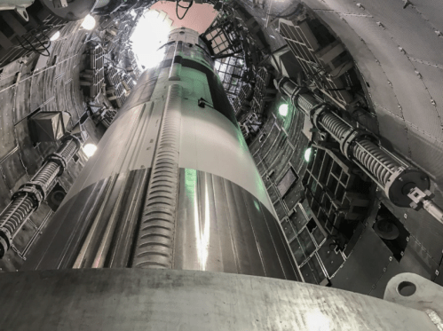 View looking up inside a cylindrical structure with metallic walls and green lights, resembling a rocket or launch silo.