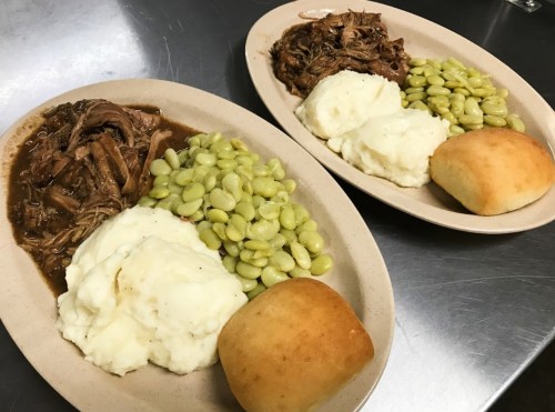 Two plates of food featuring pulled pork, mashed potatoes, lima beans, and a roll on a metal surface.
