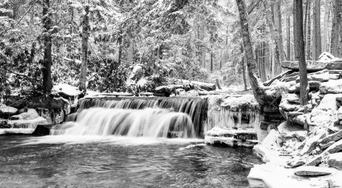 A serene black-and-white scene of a snowy forest with a cascading waterfall and still water below.