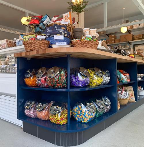A colorful display of candy jars on a wooden counter, surrounded by baskets of snacks and various packaged goods.