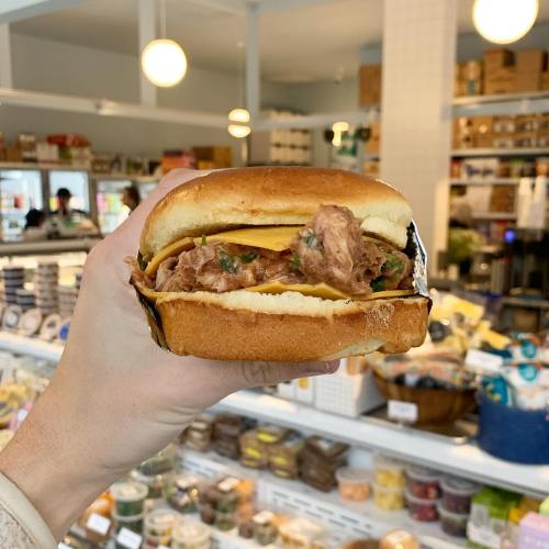 A hand holding a pulled pork sandwich with cheese, set against a background of a grocery store.