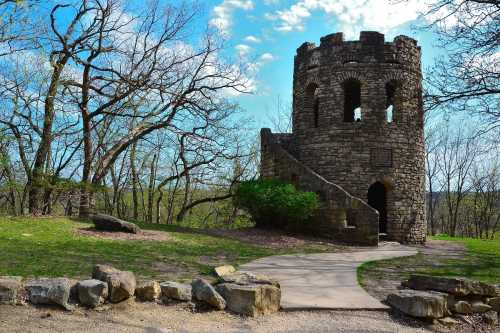 A stone tower with a staircase, surrounded by trees and a pathway, under a blue sky with scattered clouds.