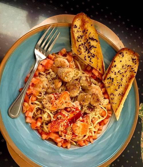 A blue plate with pasta, shrimp, lobster, diced tomatoes, and two slices of garlic bread on the side.