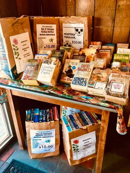 A wooden table displays various books and paper bags, featuring genres like mystery and thrillers, with prices listed.
