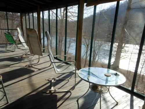 A sunlit porch with metal chairs and a glass table, overlooking a river and trees in the background.