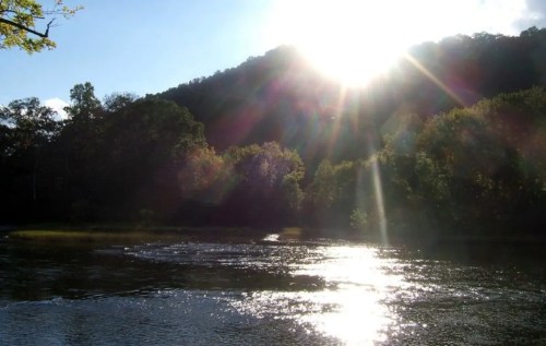Sunlight glimmers on a river, surrounded by lush trees and a mountain in the background under a clear blue sky.