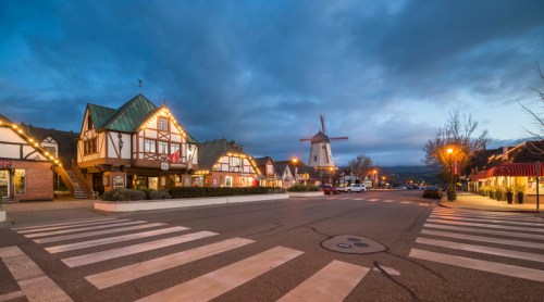 A charming street scene with Tudor-style buildings and a windmill, illuminated by streetlights at dusk.
