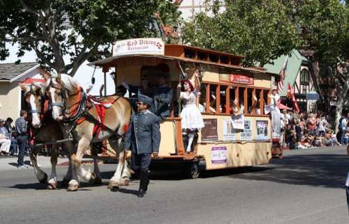 A horse-drawn trolley in a parade, with people in costumes waving to the crowd along the street.